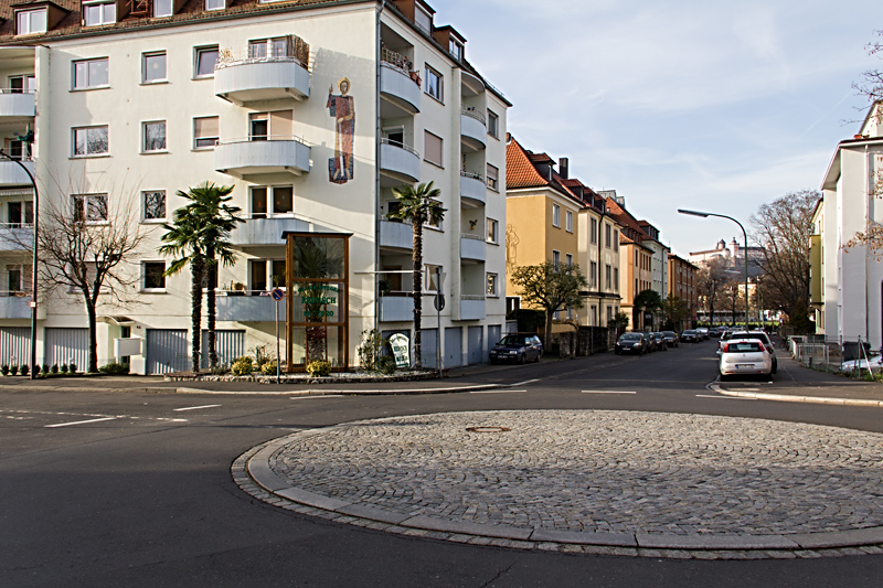 Die ehemalige Straßenkreuzung an der Neuberg- / Friedensstraße wurde vor einigen Jahren zu einem Kreisverkehr umgebaut. Vor dem Gebäude auf der linken Seite wachsen schon seit vielen Jahren die wahrscheinlich einzigen Palmen im Würzburger "Stadtgrün". Während der Wintermonate sind sie Teilweise durch eine Art Glaskasten eingehaust.